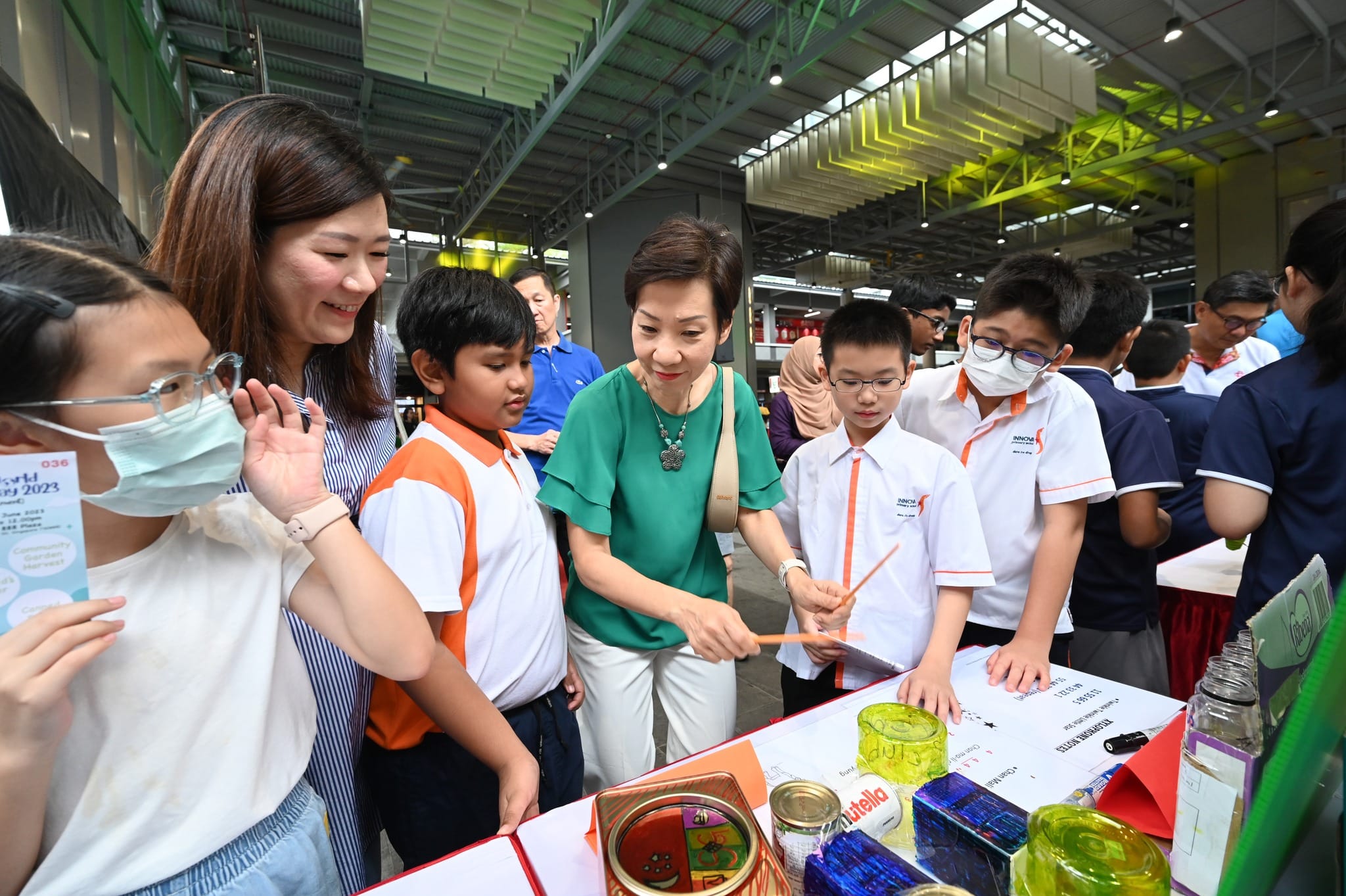 Adults and children in school uniforms gather around a display table at an indoor event, examining items together.